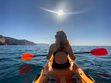 A woman in a swimsuit kayaks across a calm body of water, paddling with an orange oar. The sun shines brightly in the blue sky.
