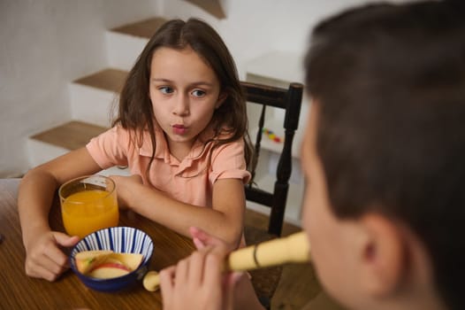 A young girl looking intrigued while sitting at a wooden table with juice and snacks, interacting with a boy playing a small musical instrument, in a casual and inviting indoor setting.