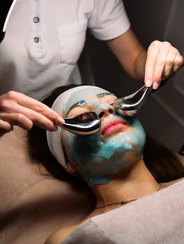 Cosmetologist does cryo massage with globules on a blue hydrogel alginate mask in the beauty salon, vertically, close-up. Woman Facial Skin & Body Care