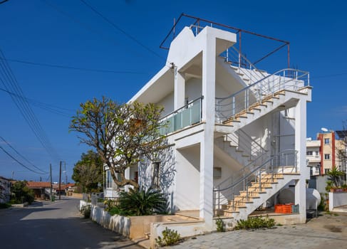 A modern building shines on a sunny street, bordered by greenery and homes beneath a clear blue sky.