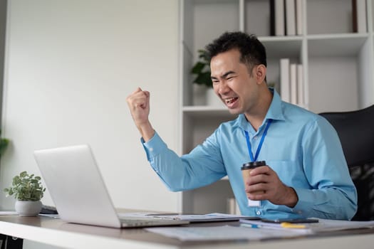 An enthusiastic Asian businessman expresses joy at his desk, celebrating a professional achievement with a coffee cup and laptop in a contemporary workspace.