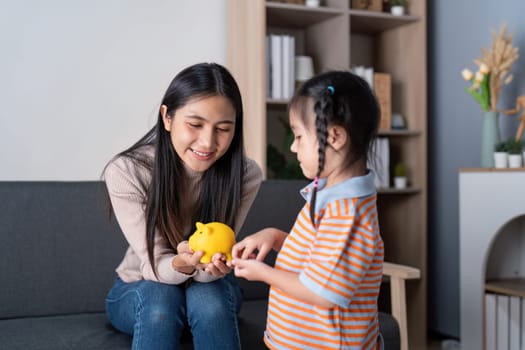 A mother joyfully teaches her daughter about saving money using a piggy bank in their inviting living room.