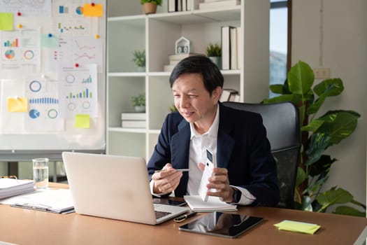 An elderly Asian businessman actively participating in a video call, taking notes while seated at his contemporary office desk.