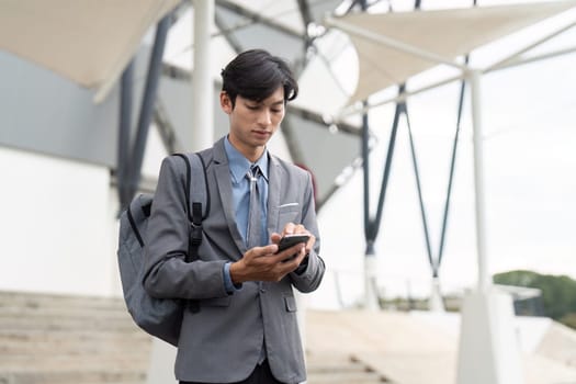 A young Asian business professional stands outside a contemporary building, engaged with his smartphone, highlighting connectivity and modern work culture.