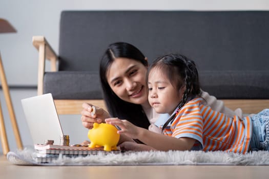 A diverse mother and her daughter enjoy a learning moment as they save money using a yellow piggy bank and a laptop, fostering financial literacy at home.