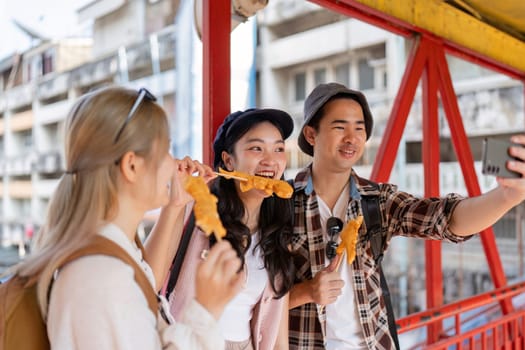 A group of young Asian friends savoring street food while capturing a fun selfie in a vibrant market, celebrating the joy of travel and camaraderie.