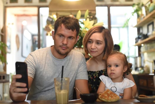 Young parents with daughter make selfie in a cafe