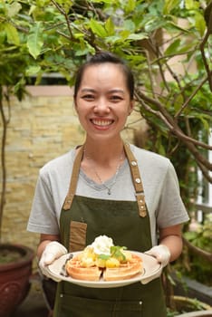 Vietnamese woman waitress serving Belgian waffles with fruits and ice cream in outdoor cafe looking up