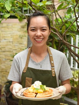 Vietnamese woman waitress serving Belgian waffles with fruits and ice cream in outdoor cafe looking up