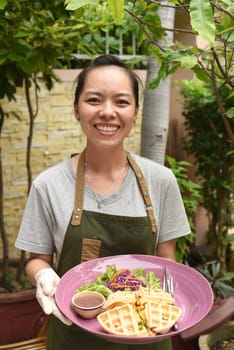 Vietnamese woman waitress serving Belgian waffles with greens and chicken in outdoor cafe