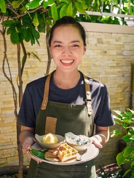 Vietnamese woman waitress serving Belgian waffles with  scoops of organic ice cream in outdoor cafe