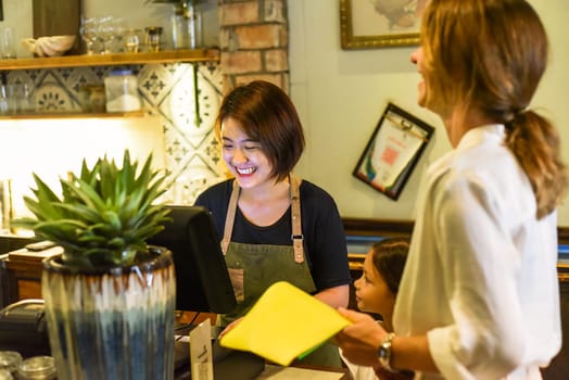 Young Vietnamese waitress working with check out machine and with customers in cafe