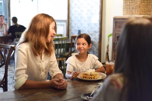 Little girl and her mother eating sweet belgian waffles with banana and cream in a cafe, daughter making a funny face