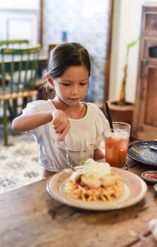 Little asian girl eating sweet belgian waffles with banana and cream in a cafe