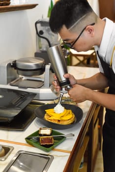 Vietnamese man chef adding fresh cream on sliced mango fruit on baked belgian waffles with cream squeezer