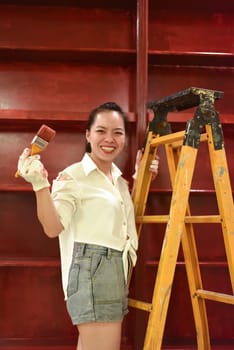 Vietnamese asian woman stands on a stepladder with a brush in her hand and paints the wall, makes repairs