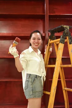 Vietnamese asian woman stands on a stepladder with a brush in her hand and paints the wall, makes repairs
