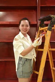 Vietnamese asian woman stands on a stepladder with a brush in her hand and paints the wall, makes repairs