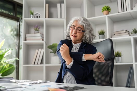 An elderly woman dressed in a business suit gently stretches her shoulder while working at her desk, focusing on self-care in her contemporary office environment.