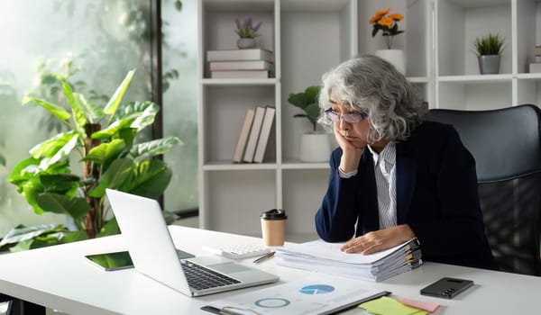 An elderly businesswoman with silver hair and glasses appears deep in thought as she reviews paperwork and her laptop in a bright, plant-filled office.