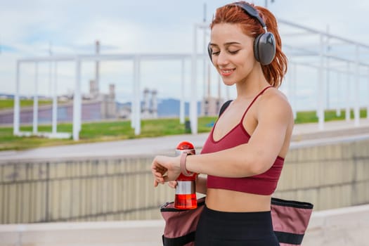Fit young woman with red hair wearing sportswear and headphones, checking her smartwatch while holding a water bottle. She carries a gym bag and stands in a modern outdoor urban environment.