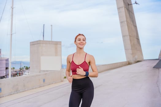 Fit young woman with red hair running outdoors near the coast. She is wearing sportswear and looks happy and energetic. The background features a bright sky and a modern urban environment.