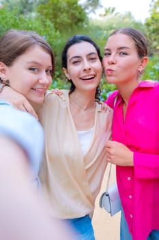 Three smiling women posing for a selfie in a lush green park. They embrace each other, expressing happiness and friendship. The vibrant colors and natural background create a joyful atmosphere.