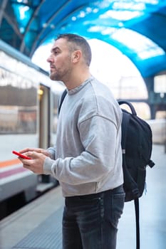 Serious man wearing a gray sweatshirt holding a red smartphone while looking around at a train station. His expression conveys anticipation, travel, and modern connectivity.