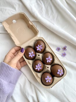 A hand holding a carton of chocolate Easter eggs decorated with fresh purple flowers, set on a white fabric background, creating a serene and festive vibe.