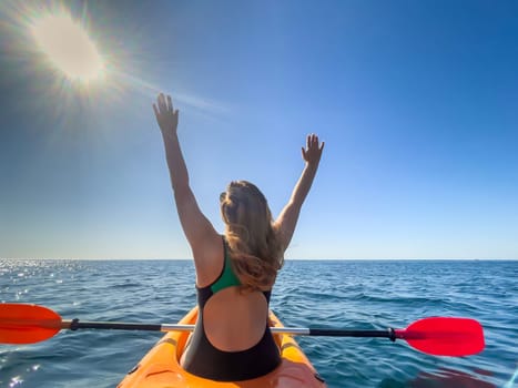 A woman in a swimsuit raises her arms in the air while kayaking on a sunny day. The ocean is calm and blue, and the sun shines brightly.