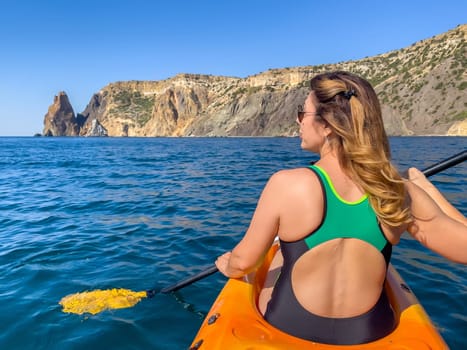A woman in a swimsuit paddles a kayak on the calm, blue water. She looks out at a rocky coastline in the distance. The sun is shining brightly.