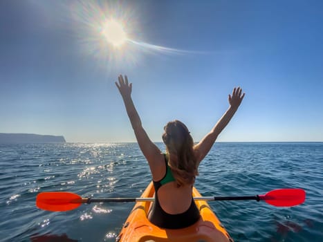 A woman in a swimsuit paddles a yellow kayak in the ocean, arms raised in the air, with the sun shining overhead.