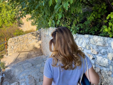 A woman with long brown hair walks up a stone staircase. Lush foliage frames the scene.