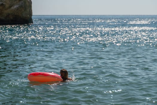 A person swims in the ocean, holding onto a bright red inflatable ring. The water is sparkling with sunlight. A rocky cliff looms in the background.