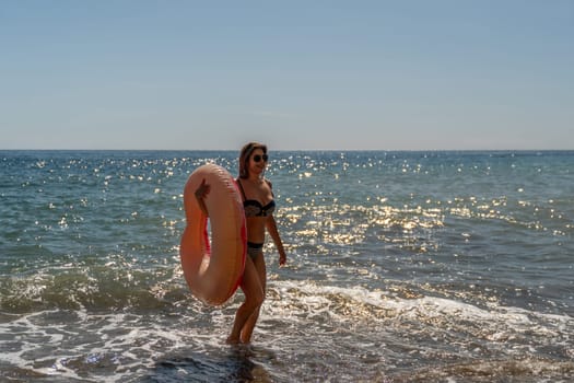 A woman in a bikini walks towards the ocean, holding a pink inflatable ring. The water is blue and sparkling, with white waves crashing on the shore.