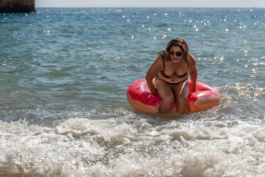A woman in a bikini sits on a pink donut pool float in the ocean. She is smiling and looks happy. The water is blue and clear. The waves are crashing on the shore.