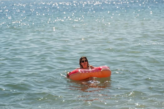 A woman floats in a bright orange inflatable ring in calm blue water, a sunny day at the beach.