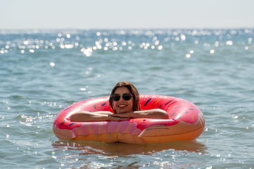 A woman wearing sunglasses floats in a pink inflatable ring in the ocean. The water is blue and the sun is shining.