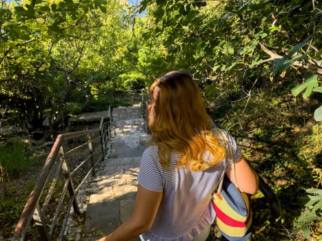 A woman with long blonde hair walks up a stone staircase with a handrail, surrounded by lush greenery. She is wearing a white t-shirt and carrying a blue and red bag.