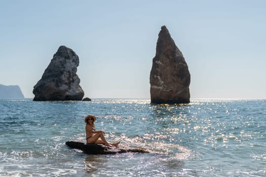 A woman sits on a rock in the water, facing two large rock formations, a calm sea stretches out behind.