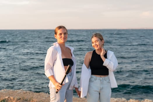 Two women stand on a rocky beach, looking at the camera. The woman on the left holds a white flute, the other woman has her hand on her cheek. The ocean is behind them.