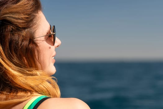 A woman with sunglasses and long hair gazes out at the ocean while wearing a swimsuit. The sun shines on the blue water.
