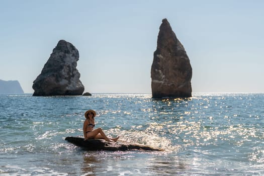 A woman sits on a rock in the ocean, facing two large rock formations in the distance. The water is blue and the sky is clear.