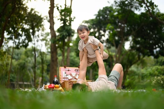 Playful father lifting his toddler into the air during a picnic in the park.