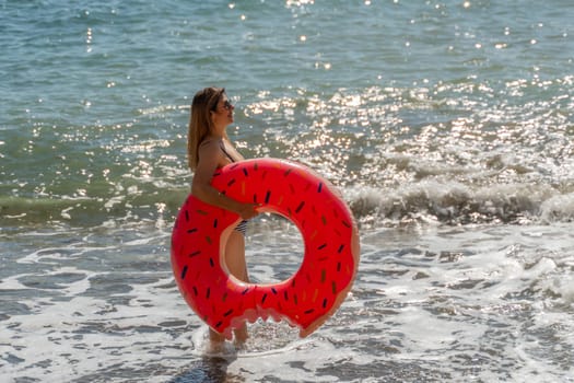 A woman in a bikini stands in the shallows of the ocean, holding a red watermelon-shaped inflatable ring.