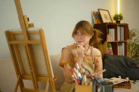 Young artist sitting on couch in studio and contemplating her artwork.