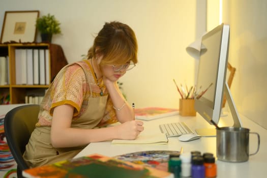 Female artist focused on sketching in a notebook, sitting at desk with computer and art supplies.