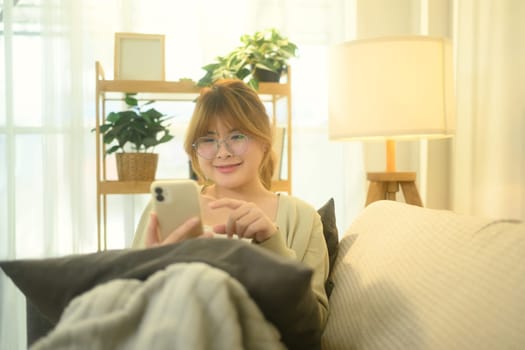 Pretty young woman in glasses texting on her mobile phone while sitting in living room.