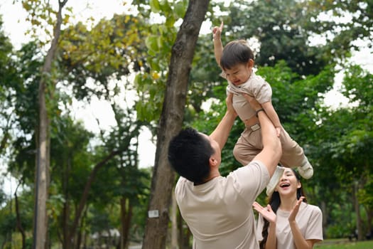 Cheerful family walking in the park, father lifting his excited toddler into the air.