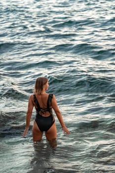 A woman in a black swimsuit stands in the ocean, facing away from the camera, with waves splashing around her.